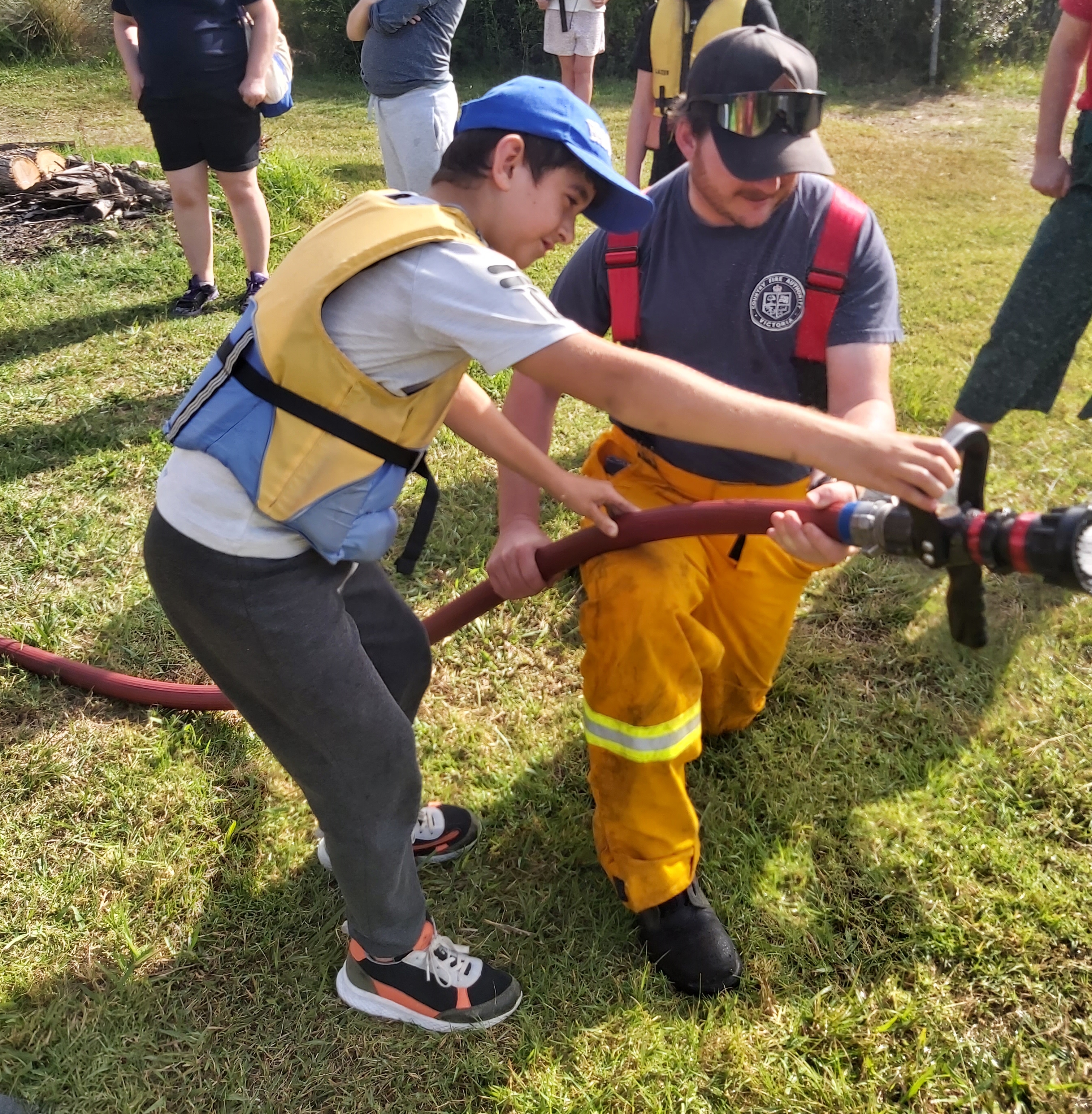 Photo of a Firefighter and a camp holding a Fire Hose. The camper is wearing a cap and a lifejacket.