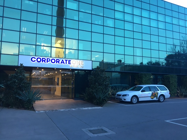 An image of the Corporate One Building. It is a 5 storey glass building. The entrance has two hedges on either side. A People Outdoors car is pictured on the right side of the entrance.