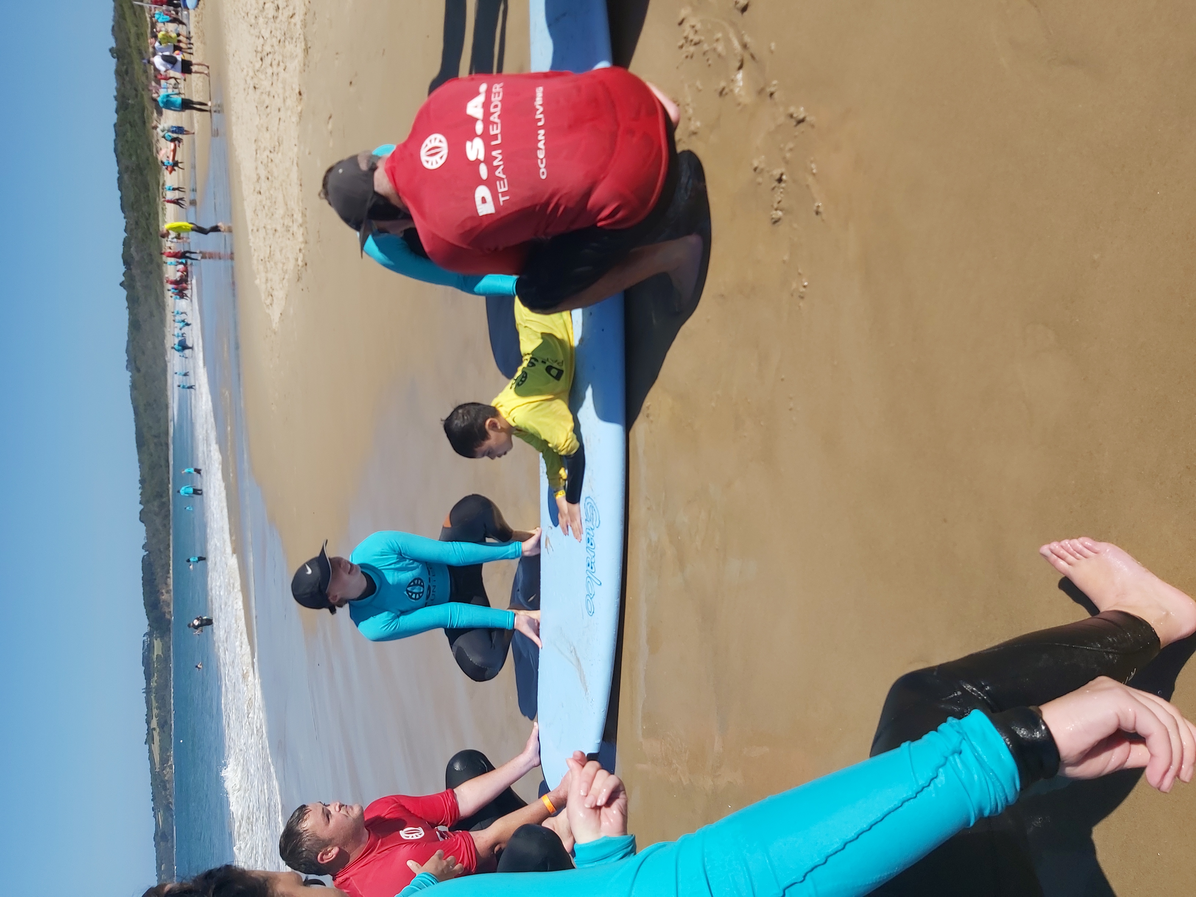 People dressed in red and blue wetsuits hold onto a surf board. A child with a yellow wetsuit and lying on the surfer. 